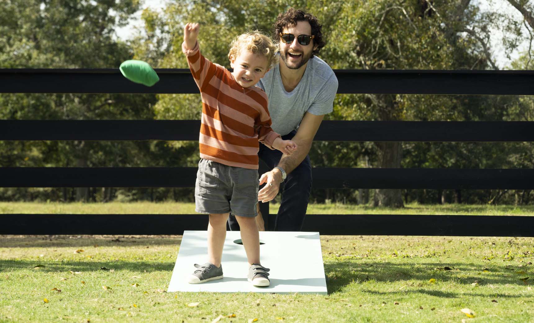 A young child with curly hair, in a striped sweater and shorts, eagerly plays Ledge Loungers Cornhole in the yard. An adult with curly hair and sunglasses, wearing a gray t-shirt, crouches beside him, smiling against a backdrop of trees and a black fence.