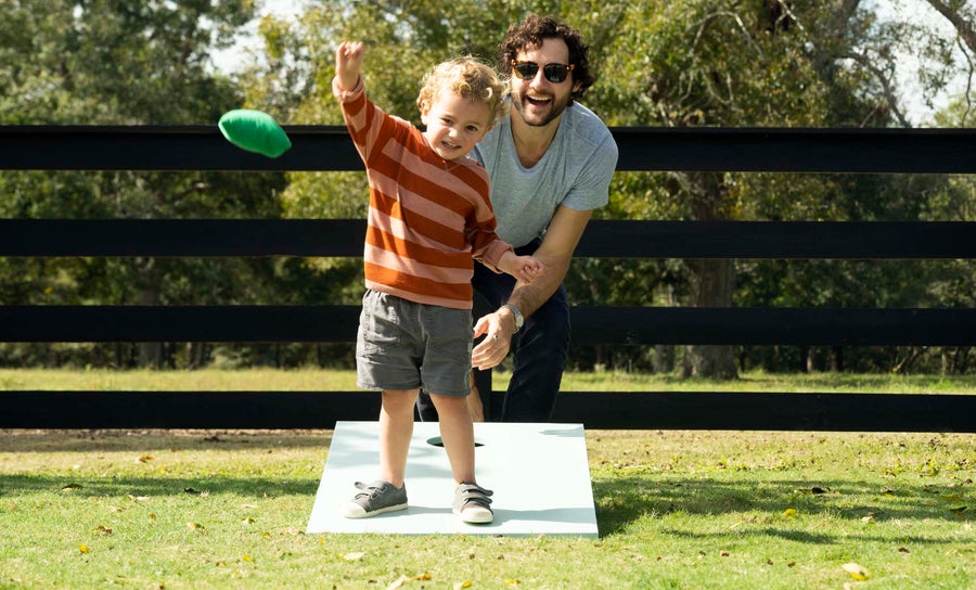 A young child with curly hair, in a striped sweater and shorts, eagerly plays Ledge Loungers Cornhole in the yard. An adult with curly hair and sunglasses, wearing a gray t-shirt, crouches beside him, smiling against a backdrop of trees and a black fence.