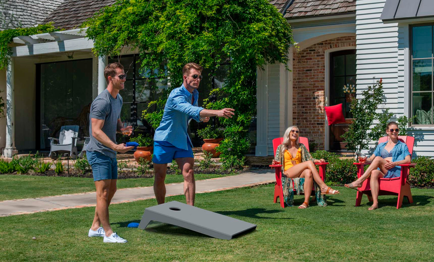 Two men participate in a Ledge Lounger Cornhole game on a lawn lined with lush greenery. One throws accurately as the other observes. Nearby, two women relax on red chairs, watching the lively scene unfold under the sun.