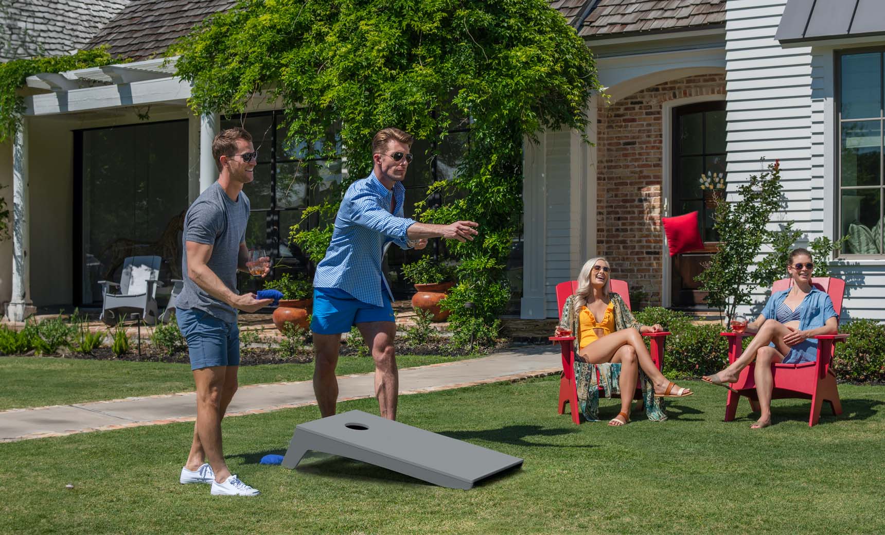 Two men participate in a Ledge Lounger Cornhole game on a lawn lined with lush greenery. One throws accurately as the other observes. Nearby, two women relax on red chairs, watching the lively scene unfold under the sun.