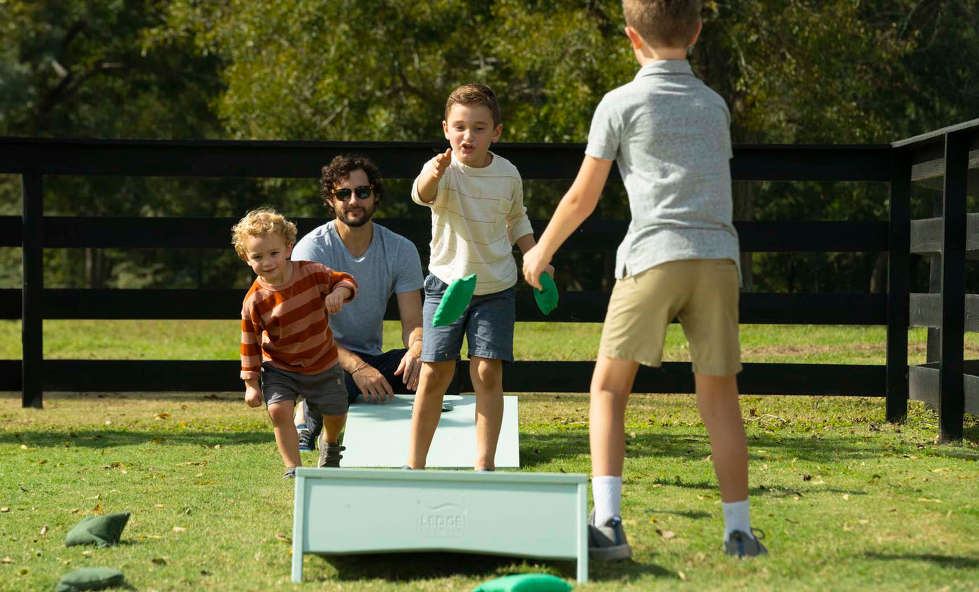A man and three kids play a lively game with the portable Ledge Lounger Cornhole set on the grass. Two children toss bean bags as another cheers. Trees and a wooden fence frame the scene beneath a clear sky.