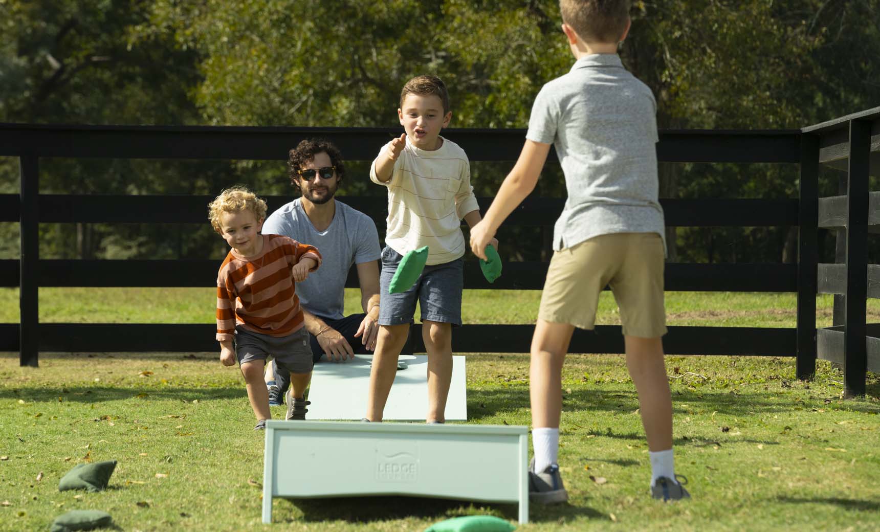 A man and three kids play a lively game with the portable Ledge Lounger Cornhole set on the grass. Two children toss bean bags as another cheers. Trees and a wooden fence frame the scene beneath a clear sky.
