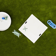 Aerial shot of the Checkers & Tic Tac Toe game by Ledge Lounger on a lawn. A white board with X and O pieces, some askew, is next to a round container full of extras. Blue rings scattered nearby enhance this classic outdoor Tic Tac Toe setup.