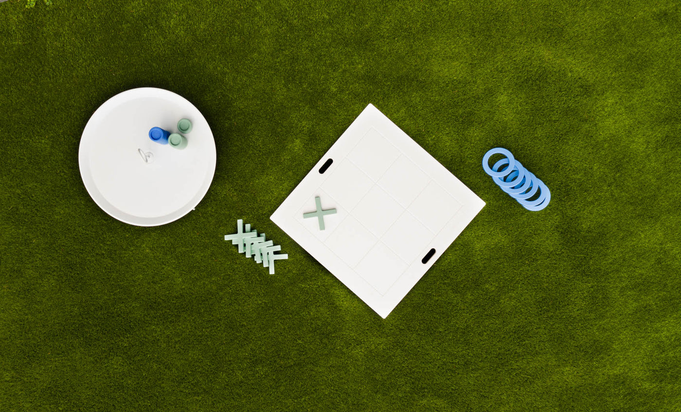 Aerial shot of the Checkers & Tic Tac Toe game by Ledge Lounger on a lawn. A white board with X and O pieces, some askew, is next to a round container full of extras. Blue rings scattered nearby enhance this classic outdoor Tic Tac Toe setup.