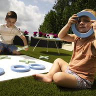 Two kids have fun with the Ledge Lounger Checkers & Tic Tac Toe game on the grass. The older one places an X as the younger playfully holds an O over their eyes. A table behind them holds more pieces amid trees and flowers.