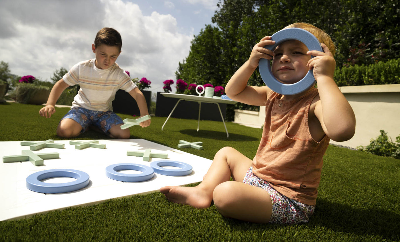 Two kids have fun with the Ledge Lounger Checkers & Tic Tac Toe game on the grass. The older one places an X as the younger playfully holds an O over their eyes. A table behind them holds more pieces amid trees and flowers.