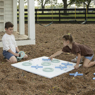Two children play with Ledge Loungers Checkers & Tic Tac Toe set on the playground amid wood chips. A boy arranges pieces while a girl kneels to place an X. In the background, a child inspects the ground, and swings gently sway nearby.