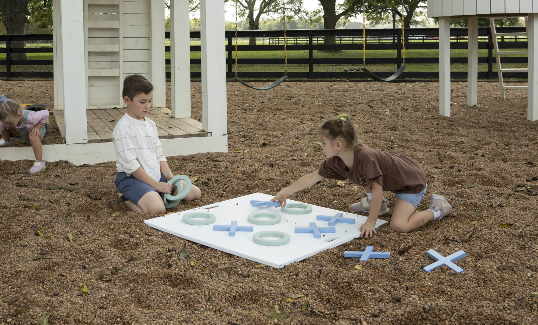 Two children play with Ledge Loungers Checkers & Tic Tac Toe set on the playground amid wood chips. A boy arranges pieces while a girl kneels to place an X. In the background, a child inspects the ground, and swings gently sway nearby.