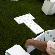 People are enjoying a lawn game with Ledge Loungers giant Dominoes on green grass. Hands are carefully arranging the pieces, which feature large green dots, into a circular pattern. The high-quality construction of Ledge Lounger Dominoes ensures long-lasting outdoor fun.