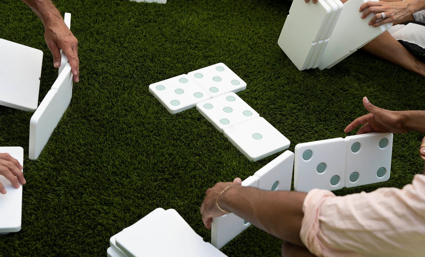 People are enjoying a lawn game with Ledge Loungers giant Dominoes on green grass. Hands are carefully arranging the pieces, which feature large green dots, into a circular pattern. The high-quality construction of Ledge Lounger Dominoes ensures long-lasting outdoor fun.