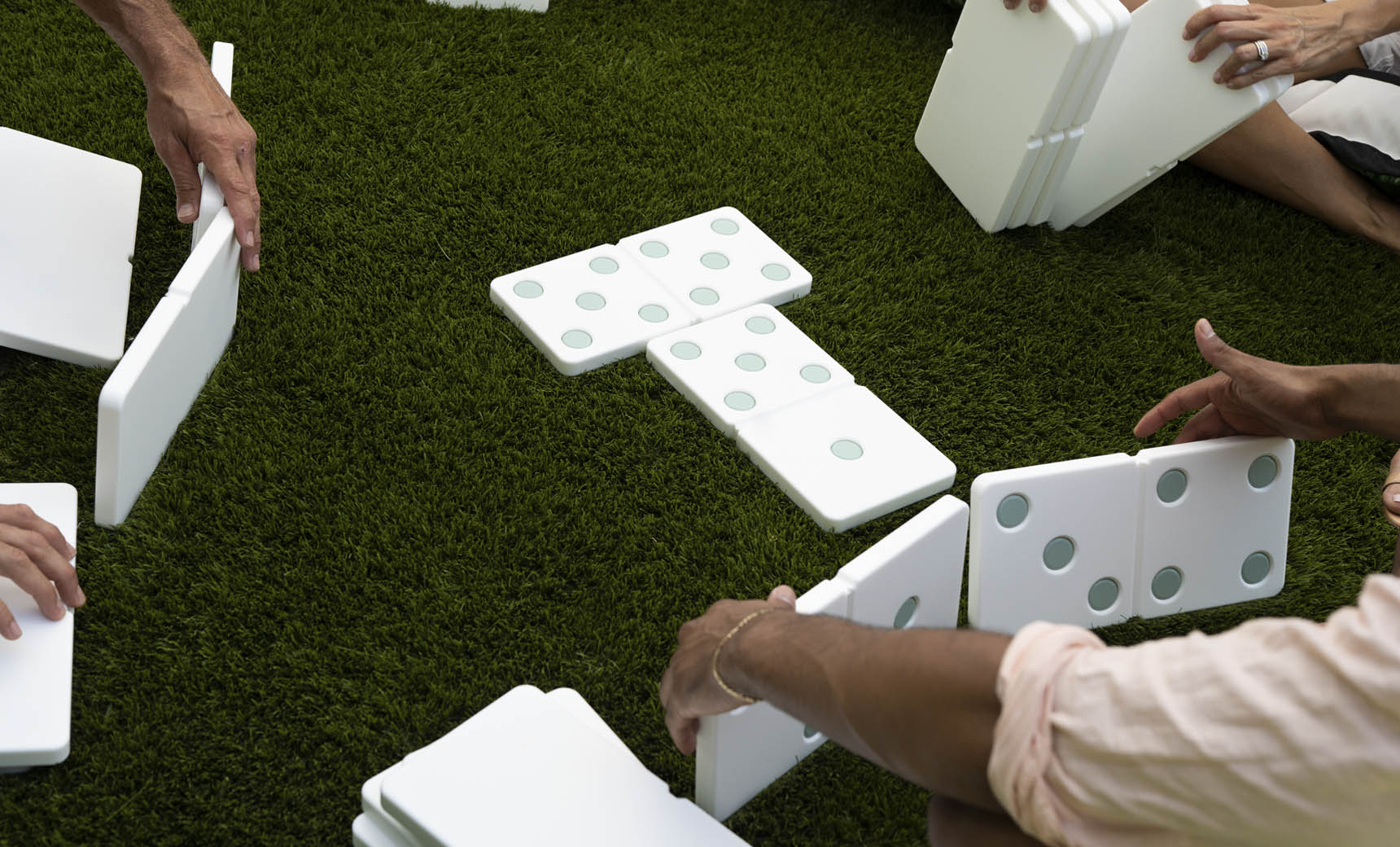 People are enjoying a lawn game with Ledge Loungers giant Dominoes on green grass. Hands are carefully arranging the pieces, which feature large green dots, into a circular pattern. The high-quality construction of Ledge Lounger Dominoes ensures long-lasting outdoor fun.