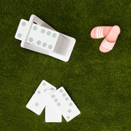 Dominoes from Ledge Lounger are scattered on the green grass, some neatly stored in a white holder. Their green dots shine under the sun near pink and white striped sandals, suggesting a joyful afternoon with a high-quality lawn game set.