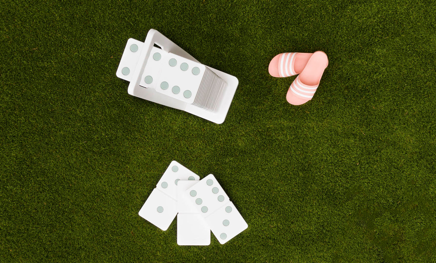 Dominoes from Ledge Lounger are scattered on the green grass, some neatly stored in a white holder. Their green dots shine under the sun near pink and white striped sandals, suggesting a joyful afternoon with a high-quality lawn game set.