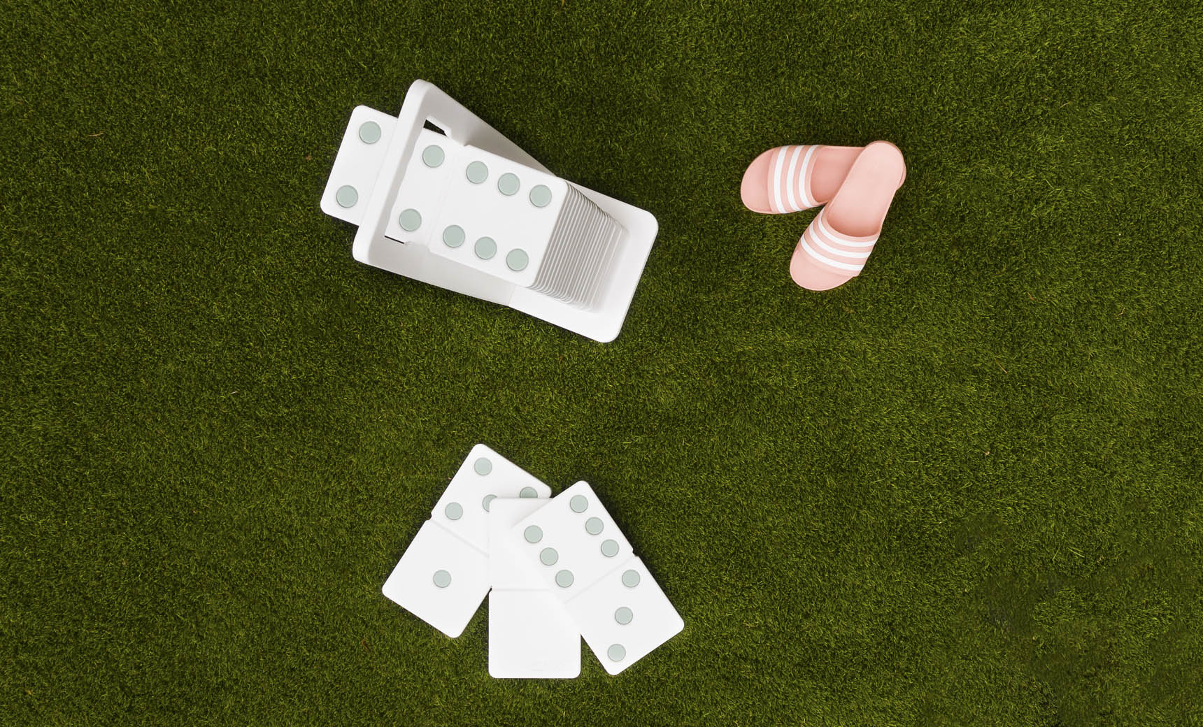 Dominoes from Ledge Lounger are scattered on the green grass, some neatly stored in a white holder. Their green dots shine under the sun near pink and white striped sandals, suggesting a joyful afternoon with a high-quality lawn game set.