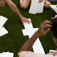 People sit on grass enjoying a lawn game with the Ledge Lounger Dominoes set. Some hold high-quality pieces, while others rest on the grass. A black and white striped cushion adds to the relaxed atmosphere in the background.