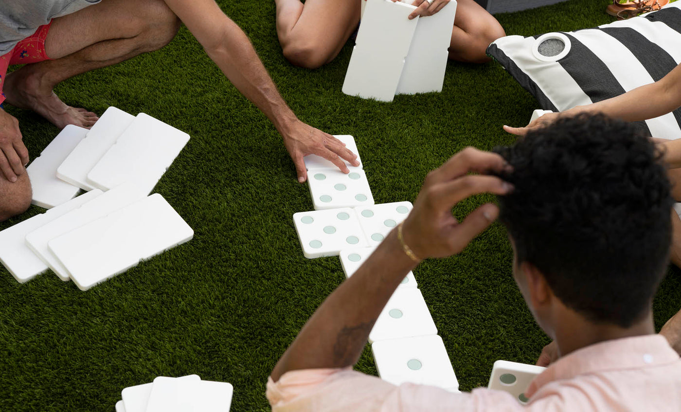 People sit on grass enjoying a lawn game with the Ledge Lounger Dominoes set. Some hold high-quality pieces, while others rest on the grass. A black and white striped cushion adds to the relaxed atmosphere in the background.