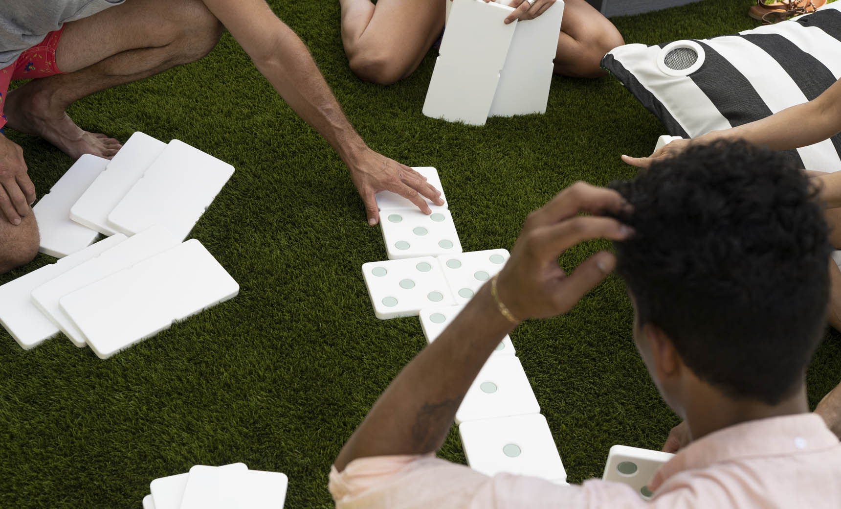 People sit on grass enjoying a lawn game with the Ledge Lounger Dominoes set. Some hold high-quality pieces, while others rest on the grass. A black and white striped cushion adds to the relaxed atmosphere in the background.