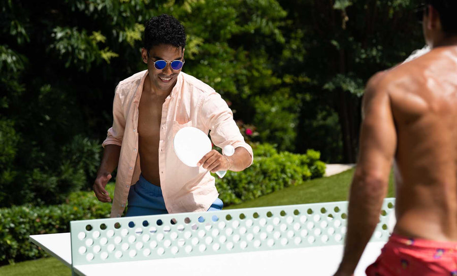 A man in a light pink shirt and sunglasses enjoys outdoor entertainment, playing ping pong with a casually dressed person. They play on a Ledge Lounger Table Tennis Set, featuring a distinct hole pattern on the net, surrounded by lush green foliage.