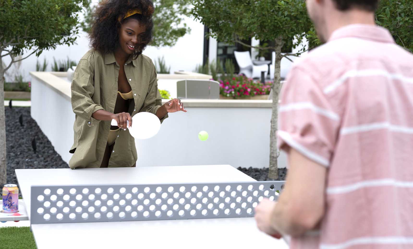 A curly-haired woman in a green jacket plays outdoor ping pong with a man in a pink shirt using the Ledge Lounger Table Tennis Set, amidst trees and flowers, ready to hit the ball. A can rests on the table nearby.