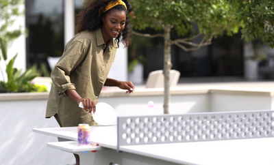 A smiling woman with curly hair and a yellow headband plays table tennis outdoors, holding a paddle and watching the ball mid-air over the table. A drink sits nearby, and greenery is in the background.