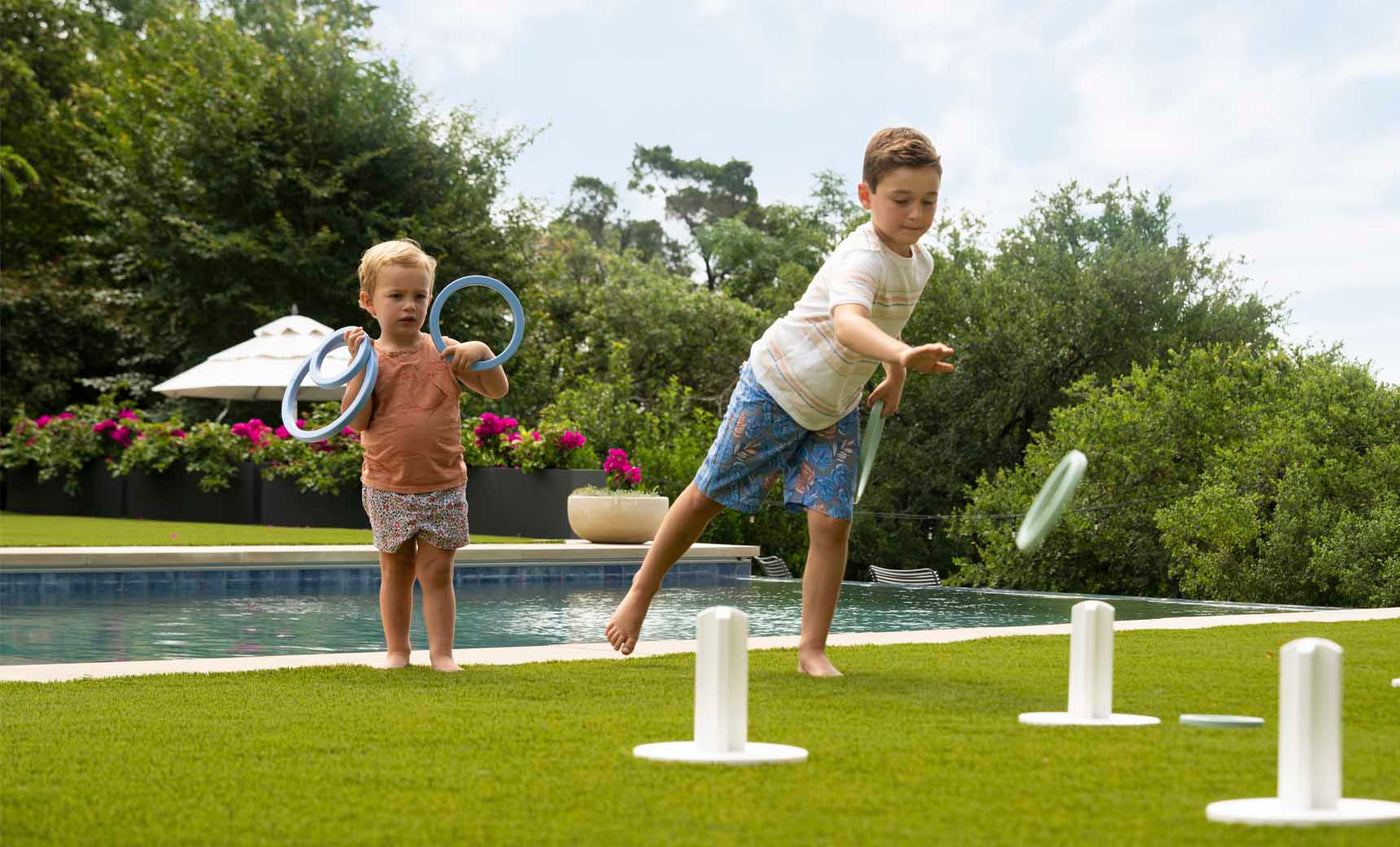 Two children play Ledge Loungers Ring Toss on a grassy lawn by a pool. The older child aims for the stakes as the younger one watches, holding rings. Trees and flowers frame this lively outdoor scene.