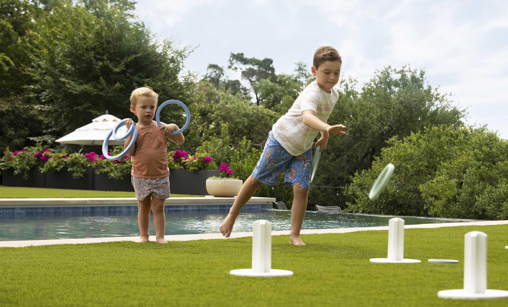 Two children play Ledge Loungers Ring Toss on a grassy lawn by a pool. The older child aims for the stakes as the younger one watches, holding rings. Trees and flowers frame this lively outdoor scene.
