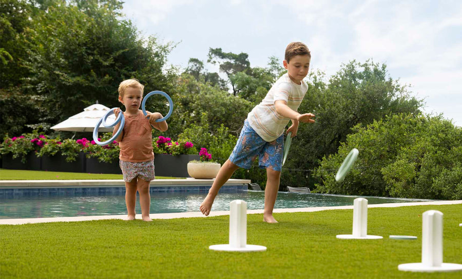 Two children play Ledge Loungers Ring Toss on a grassy lawn by a pool. The older child aims for the stakes as the younger one watches, holding rings. Trees and flowers frame this lively outdoor scene.