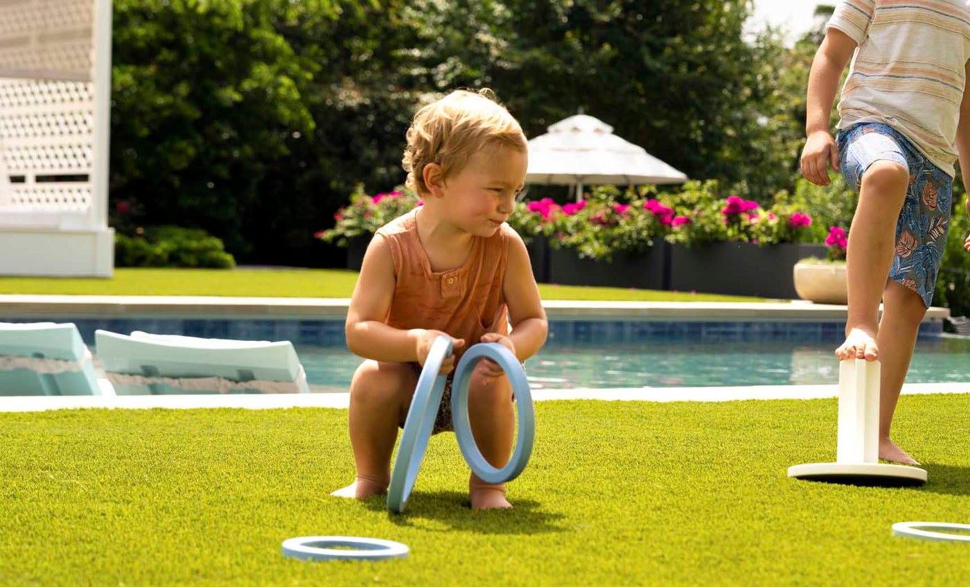 A toddler crouches on lush grass, enjoying the Ledge Lounger Ring Toss with a plastic ring by the pool. Another child balances on a circular object. Bright flowers and patio furniture add to the backyard fun in the sunny setting.