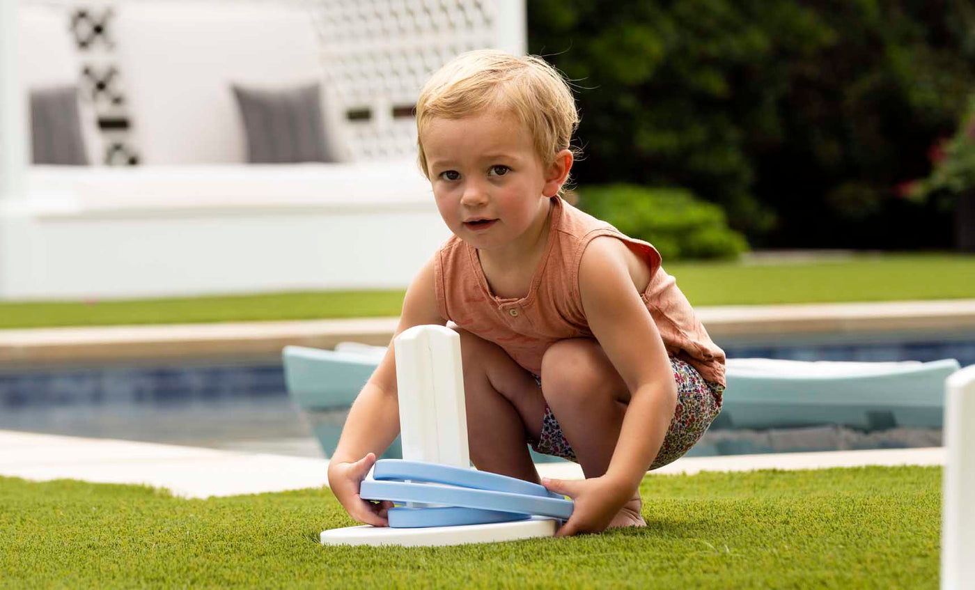 A young child with short blonde hair crouches on grass, playing Ledge Loungers Ring Toss using a blue and white plastic ring. Behind them is a pool and a cushioned seating area amidst greenery. The child sports a sleeveless orange top and patterned shorts—ideal for backyard fun.