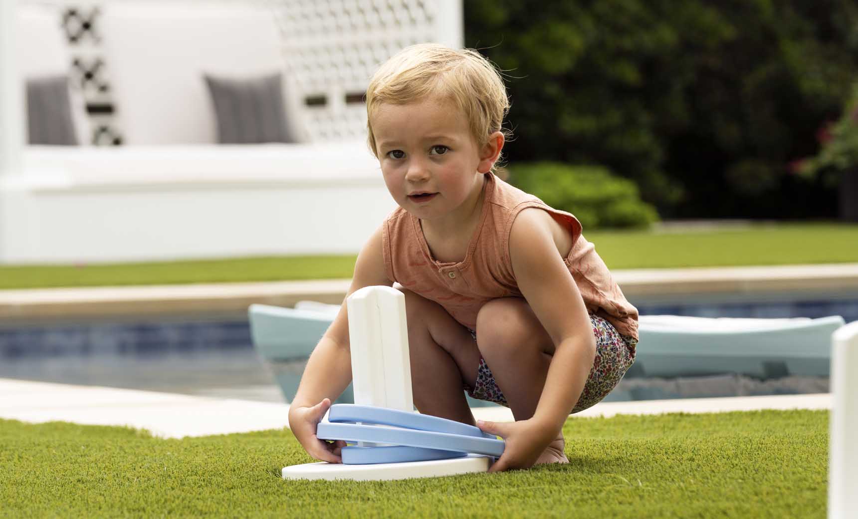 A young child with short blonde hair crouches on grass, playing Ledge Loungers Ring Toss using a blue and white plastic ring. Behind them is a pool and a cushioned seating area amidst greenery. The child sports a sleeveless orange top and patterned shorts—ideal for backyard fun.
