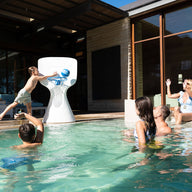 A child leaps off the pools edge, skillfully dunking a basketball into the large Ledge Lounger Hoopstr. Around him, kids and two adults cheer in a lively backyard with modern patio furniture, epitomizing summer fun.