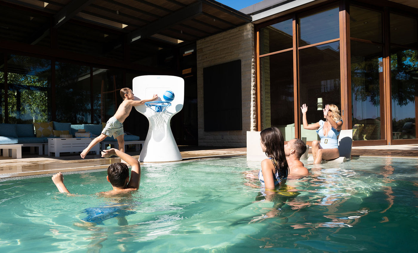 A child leaps off the pools edge, skillfully dunking a basketball into the large Ledge Lounger Hoopstr. Around him, kids and two adults cheer in a lively backyard with modern patio furniture, epitomizing summer fun.