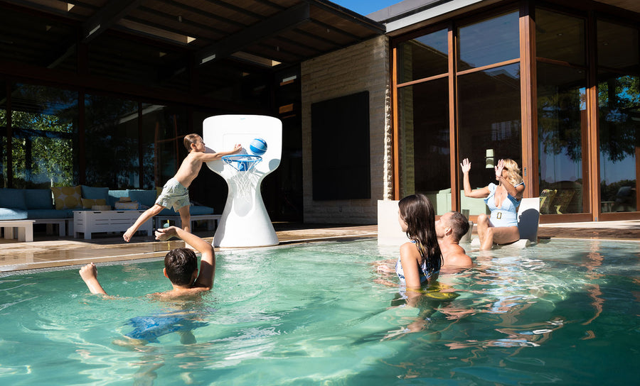 A child leaps off the pools edge, skillfully dunking a basketball into the large Ledge Lounger Hoopstr. Around him, kids and two adults cheer in a lively backyard with modern patio furniture, epitomizing summer fun.