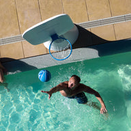A man in swim trunks enthusiastically plays backyard basketball, aiming an aqua-blue ball at a Ledge Loungers Hoopstr pool hoop. Meanwhile, a woman in a yellow swimsuit observes from the sunlit tiled deck by the poolside as clear water sparkles around them.