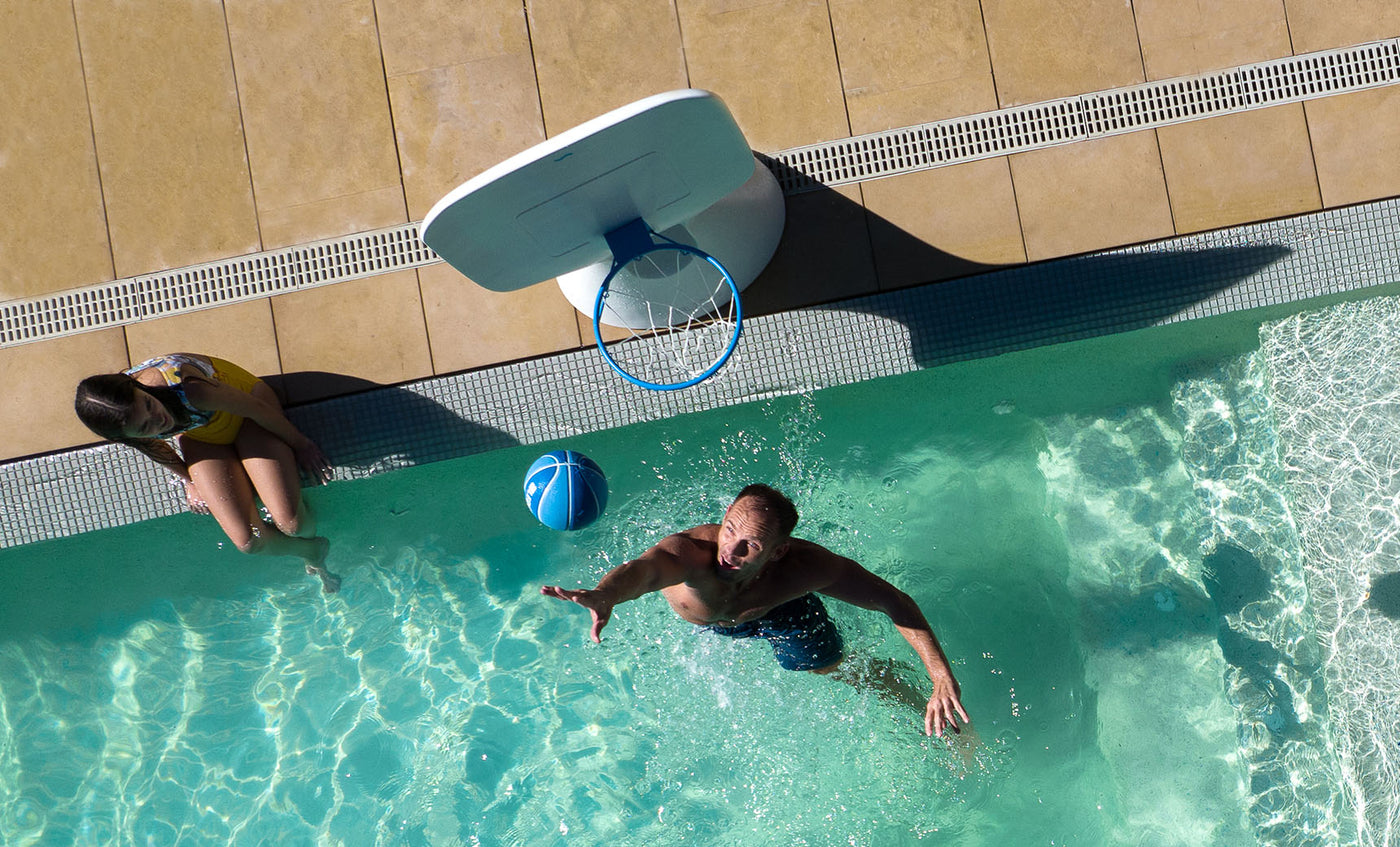 A man in swim trunks enthusiastically plays backyard basketball, aiming an aqua-blue ball at a Ledge Loungers Hoopstr pool hoop. Meanwhile, a woman in a yellow swimsuit observes from the sunlit tiled deck by the poolside as clear water sparkles around them.