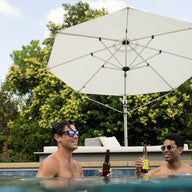 Two people in a pool hold beer bottles and smile at each other, both wearing sunglasses. Behind them, the Ledge Lounger Ultra Cantilever Umbrella - 11 Octagon provides shade over a patio with a lounge chair, set against a backdrop of lush green trees.