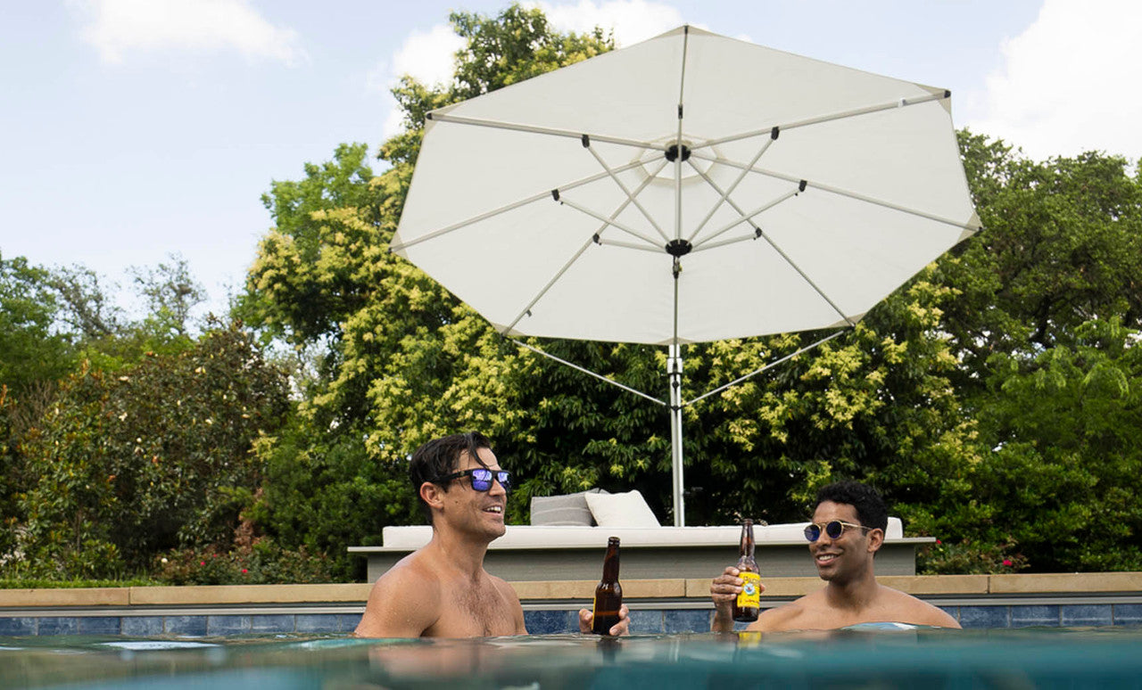 Two people in a pool hold beer bottles and smile at each other, both wearing sunglasses. Behind them, the Ledge Lounger Ultra Cantilever Umbrella - 11 Octagon provides shade over a patio with a lounge chair, set against a backdrop of lush green trees.