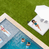 An aerial view features a pool scene: two people relax on floating loungers, while two others swim beneath the bright summer sky. A woman lounges under the 11 Octagon Ultra Cantilever Umbrella by Ledge Lounger, adding a soft glow and enhancing the inviting patio space.