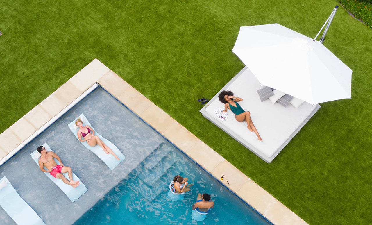 An aerial view features a pool scene: two people relax on floating loungers, while two others swim beneath the bright summer sky. A woman lounges under the 11 Octagon Ultra Cantilever Umbrella by Ledge Lounger, adding a soft glow and enhancing the inviting patio space.