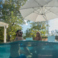 A man and woman relax in a sunny, partially submerged pool with clear water under the Ledge Lounger Ultra Cantilever Umbrella - 10 Square, enhanced by stylish patio lighting. Surrounding trees and patio furniture complete the serene scene.