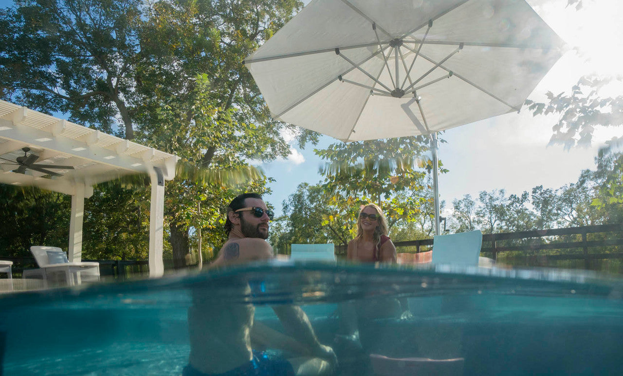A man and woman relax in a sunny, partially submerged pool with clear water under the Ledge Lounger Ultra Cantilever Umbrella - 10 Square, enhanced by stylish patio lighting. Surrounding trees and patio furniture complete the serene scene.