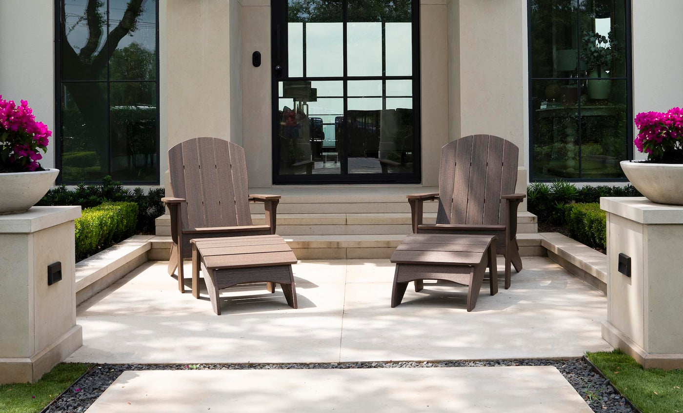 Two Legacy Adirondack by Ledge Lounger chairs with footrests sit on a patio in front of a modern building, ideal for gatherings. Behind them are large windows and stairs, while planters with purple flowers and trimmed hedges add to the inviting outdoor setup.