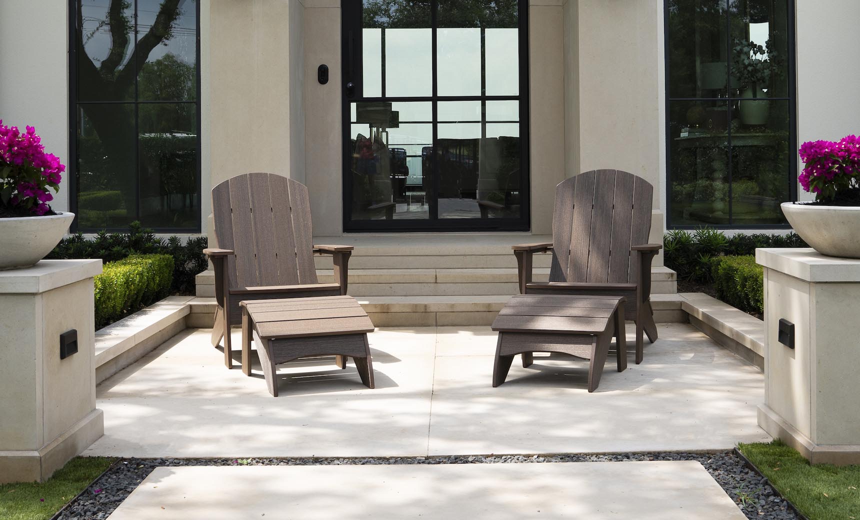 Two Legacy Adirondack by Ledge Lounger chairs with footrests sit on a patio in front of a modern building, ideal for gatherings. Behind them are large windows and stairs, while planters with purple flowers and trimmed hedges add to the inviting outdoor setup.