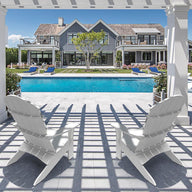 Two Legacy Adirondack chairs by Ledge Lounger sit under a pergola facing an elegant pool, with a luxurious house surrounded by greenery in the background. Sunlight casts gentle shadows on the stone patio, setting a perfect scene for intimate gatherings.