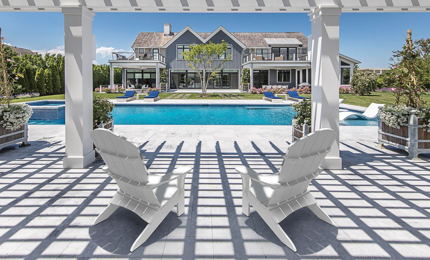 Two Legacy Adirondack chairs by Ledge Lounger sit under a pergola facing an elegant pool, with a luxurious house surrounded by greenery in the background. Sunlight casts gentle shadows on the stone patio, setting a perfect scene for intimate gatherings.