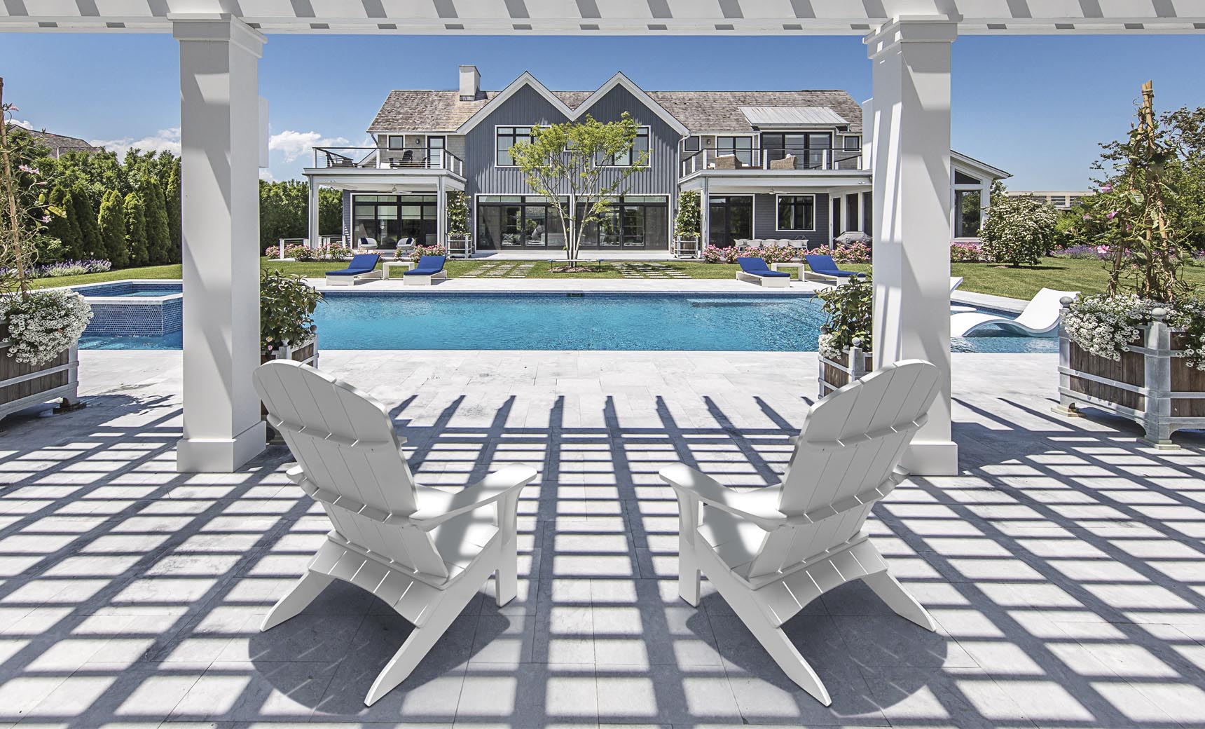 Two Legacy Adirondack chairs by Ledge Lounger sit under a pergola facing an elegant pool, with a luxurious house surrounded by greenery in the background. Sunlight casts gentle shadows on the stone patio, setting a perfect scene for intimate gatherings.