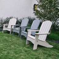 Arranged on a grassy lawn, four Legacy Adirondack chairs by Ledge Lounger (two light gray and two dark gray) create an inviting outdoor space. They face away from a white building with black-trimmed windows, near green shrubbery and a small tree.