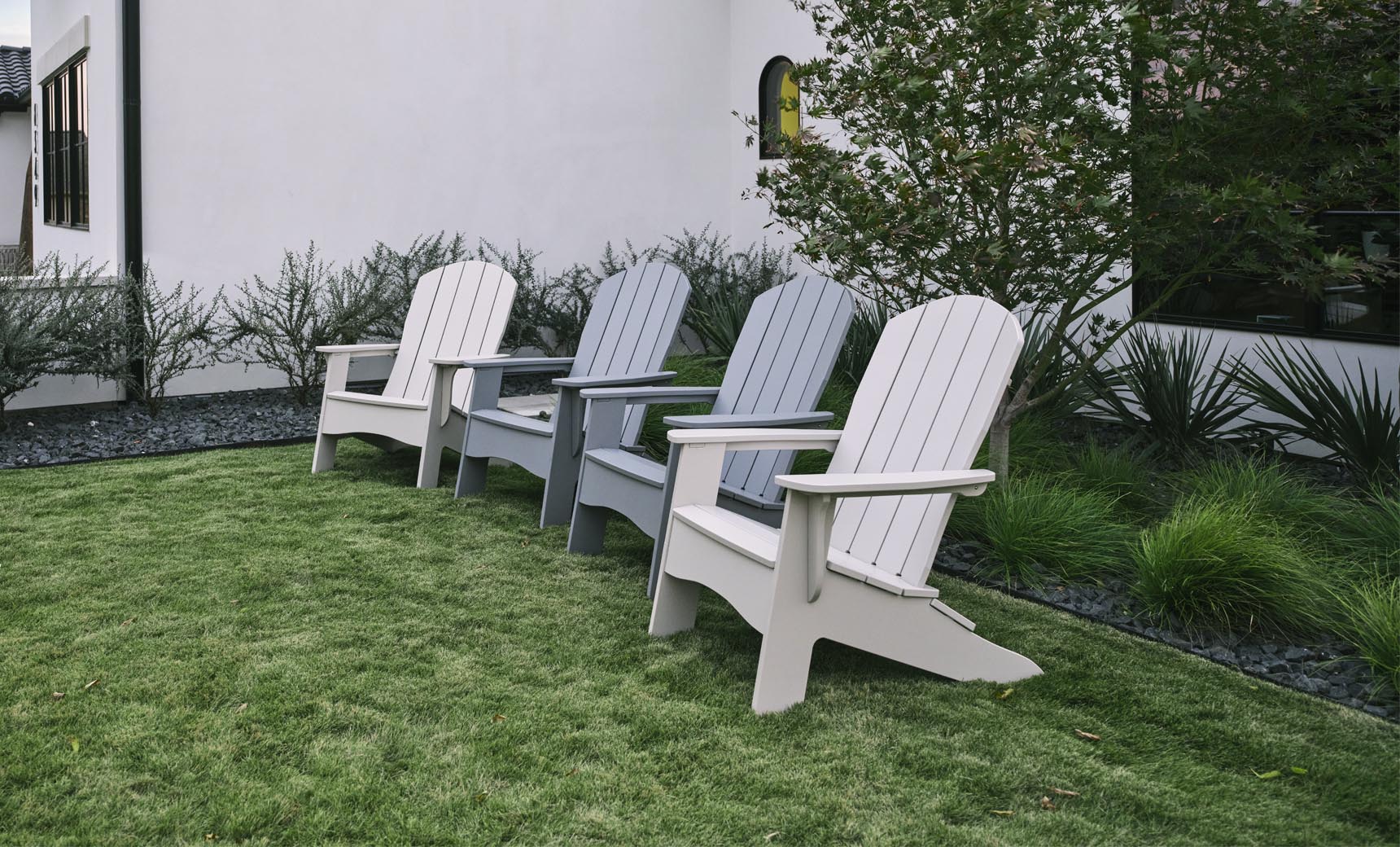 Arranged on a grassy lawn, four Legacy Adirondack chairs by Ledge Lounger (two light gray and two dark gray) create an inviting outdoor space. They face away from a white building with black-trimmed windows, near green shrubbery and a small tree.