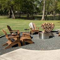 A circular outdoor seating area features Legacy Adirondack chairs by Ledge Lounger around a metal fire pit, ideal for patio gatherings. Firewood is stacked nearby, surrounded by grass and trees with a serene pond in the background on this sunny day.