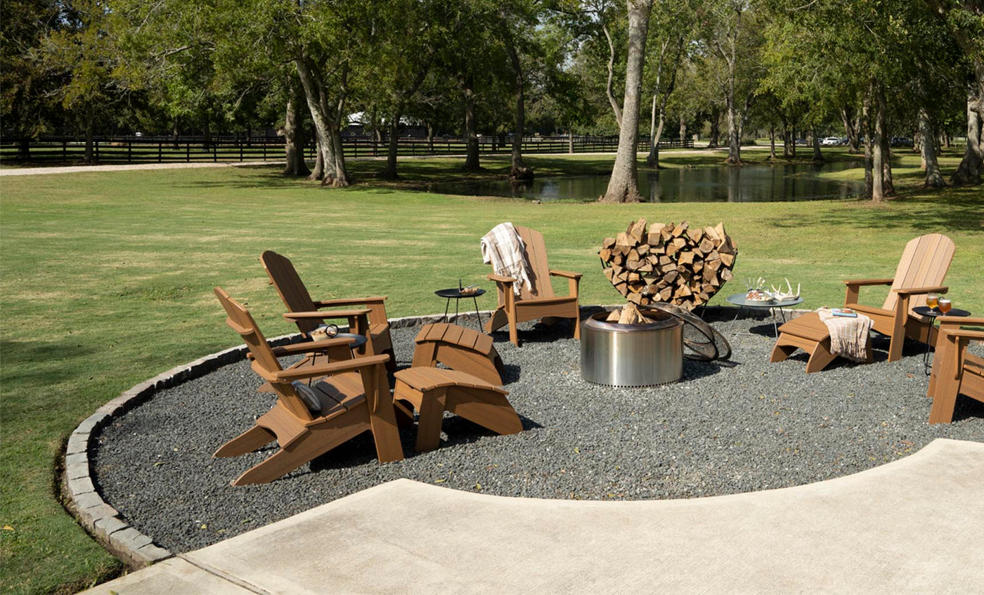 A circular outdoor seating area features Legacy Adirondack chairs by Ledge Lounger around a metal fire pit, ideal for patio gatherings. Firewood is stacked nearby, surrounded by grass and trees with a serene pond in the background on this sunny day.